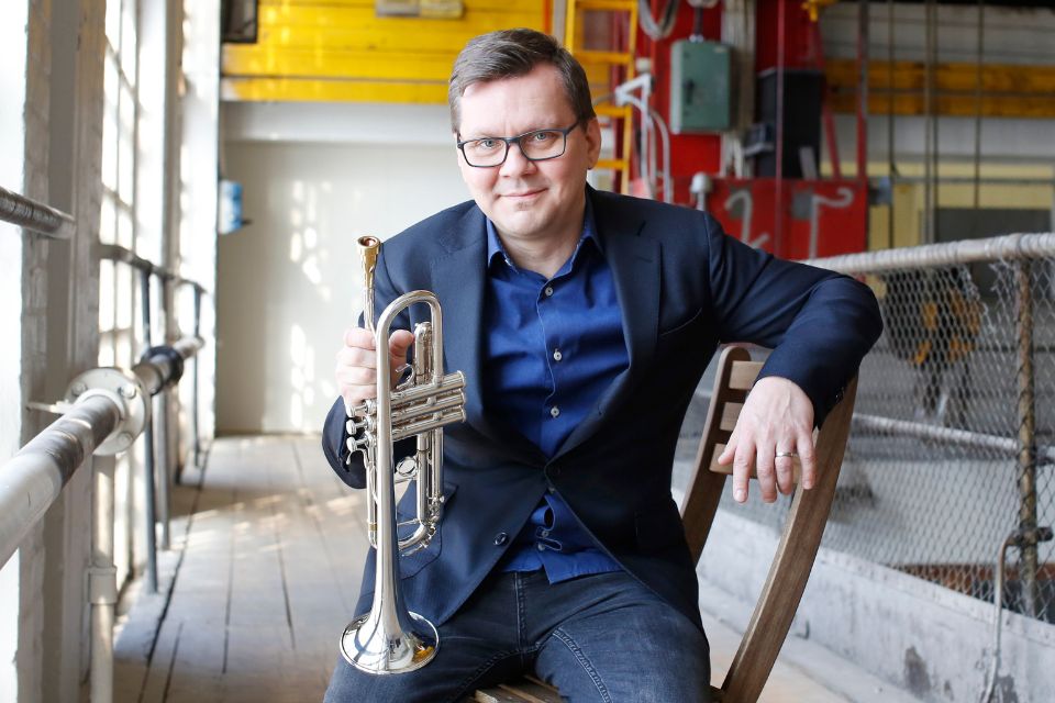Pasi Pirinen sits on a chair holding his trumpet against a colourful background