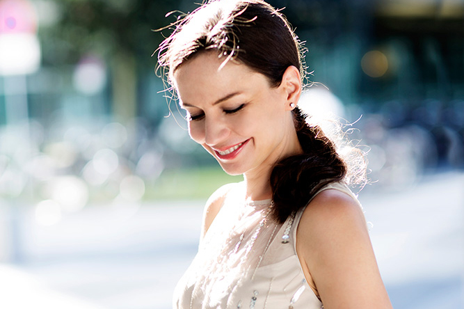 A woman with dark hair and a light dress standing outside, looking down and smiling