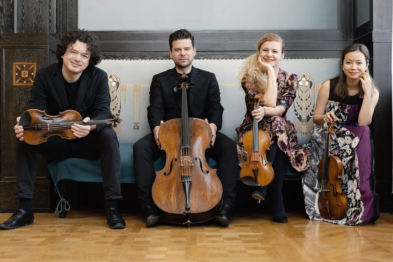 Pavel Haas Quartet sitting on a bench inside an ornate room, holding their instruments and smiling at the camera