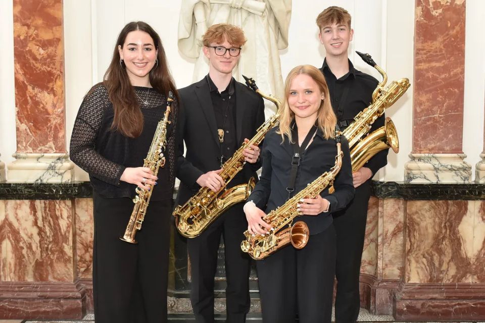 Kezia Colton and the RCMJD Saxophone Quartet wear black and hold their saxophones, smiling
