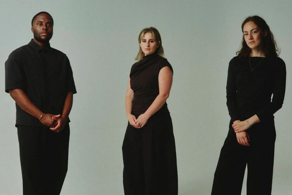 Three people wearing black stand in a brightly lit room