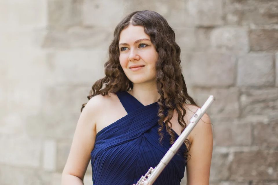 A young woman wearing a blue dress holds a flute in front of a stone wall
