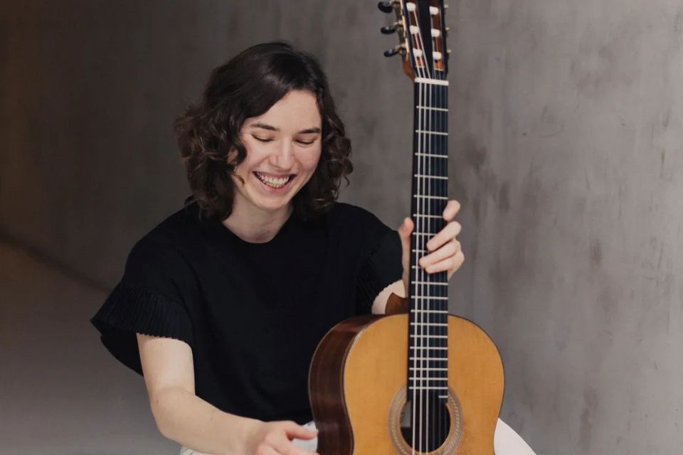 A young woman wearing black holds a guitar and smiles