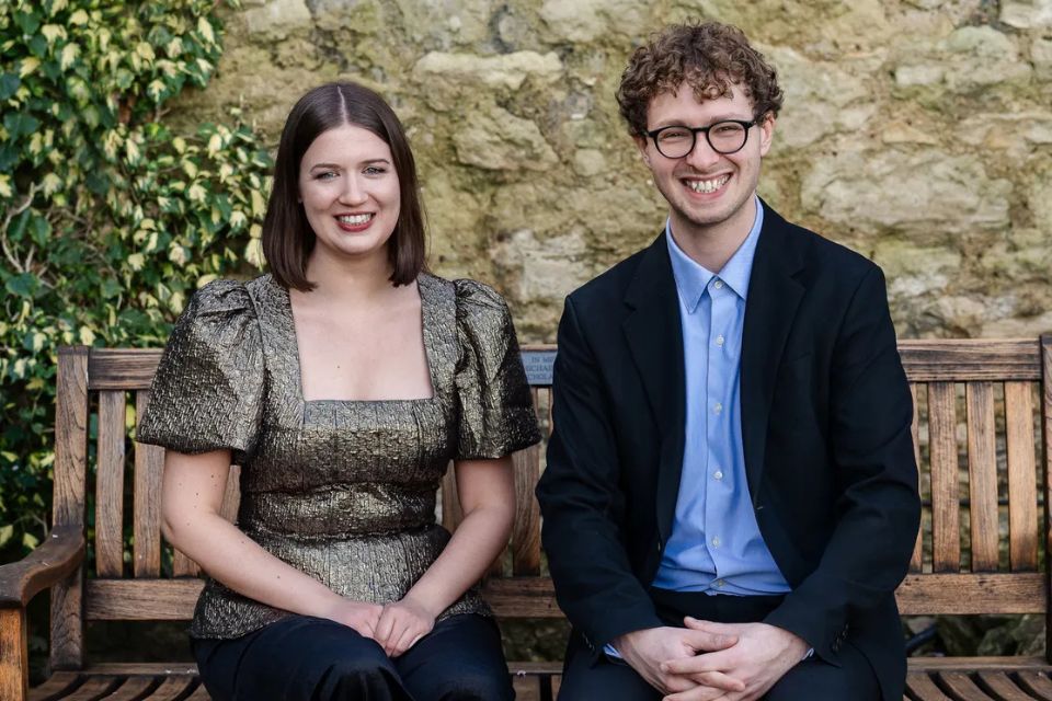 A young woman and man sit on a bench outside and smile