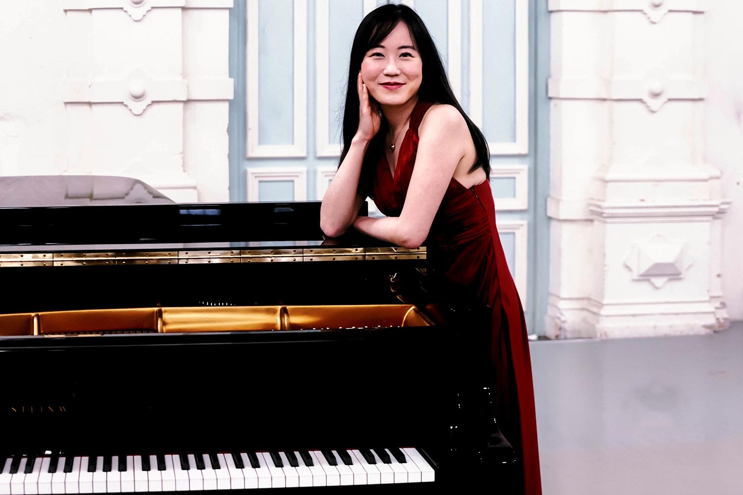 A woman wearing a dark red dress leans against a piano and smiles at the camera