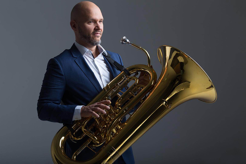 Standing against a plain grey background, tuba player Roland Szentpali holds his instrument