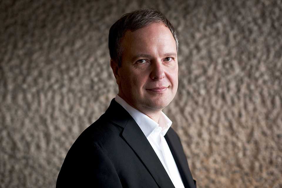 Headshot of conductor Sakari Oramo, wearing a shirt and jacket in front of a concrete wall. 