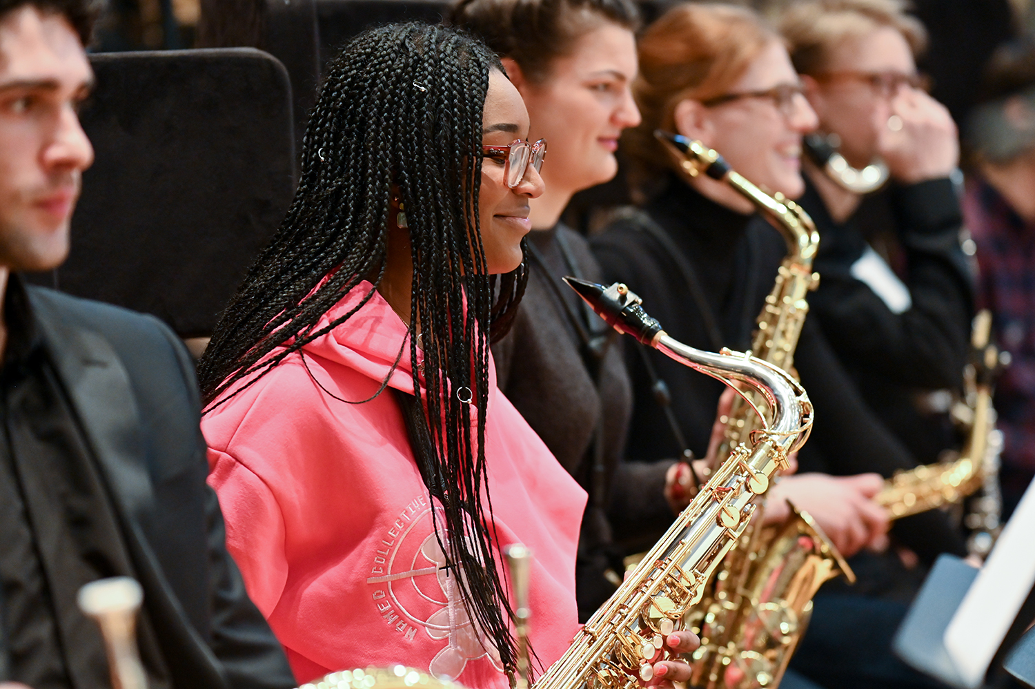 A row of saxophonists perform with the focus on one woman wearing a pink jumper