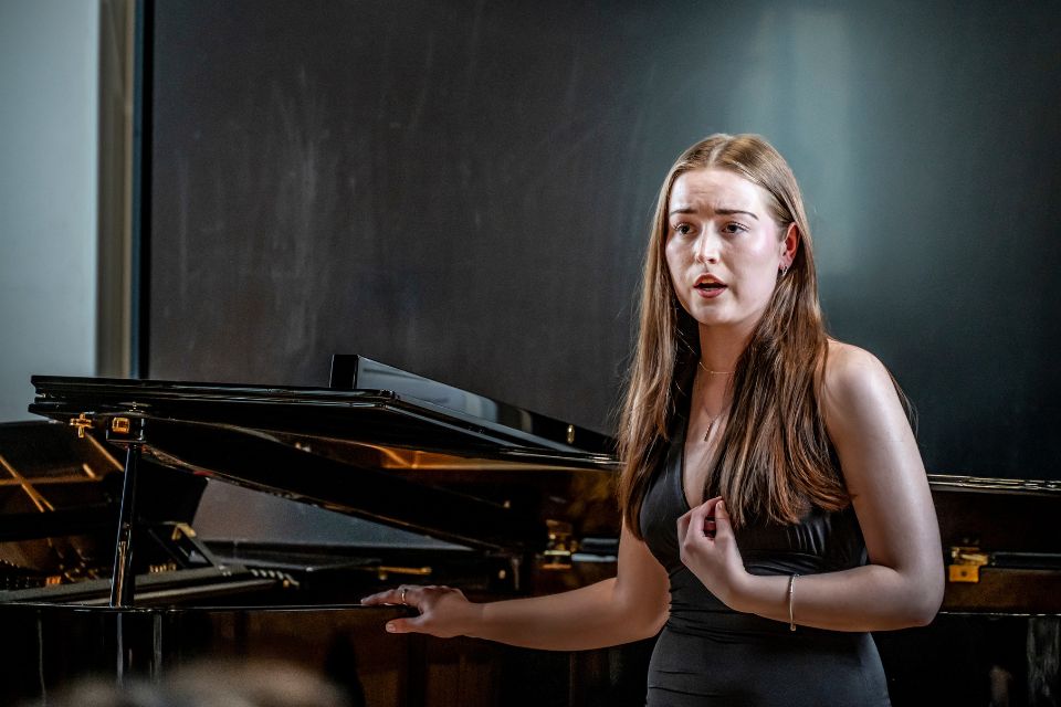 A young woman rests a hand on a piano behind her and sings passionately