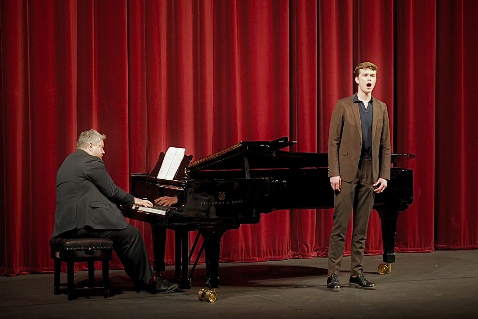 A singer performs on stage with a pianist in front of a red curtain