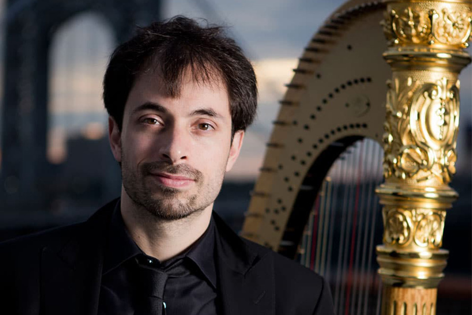 Sivan Magen, wearing black, stands next to his harp and smiles to camera