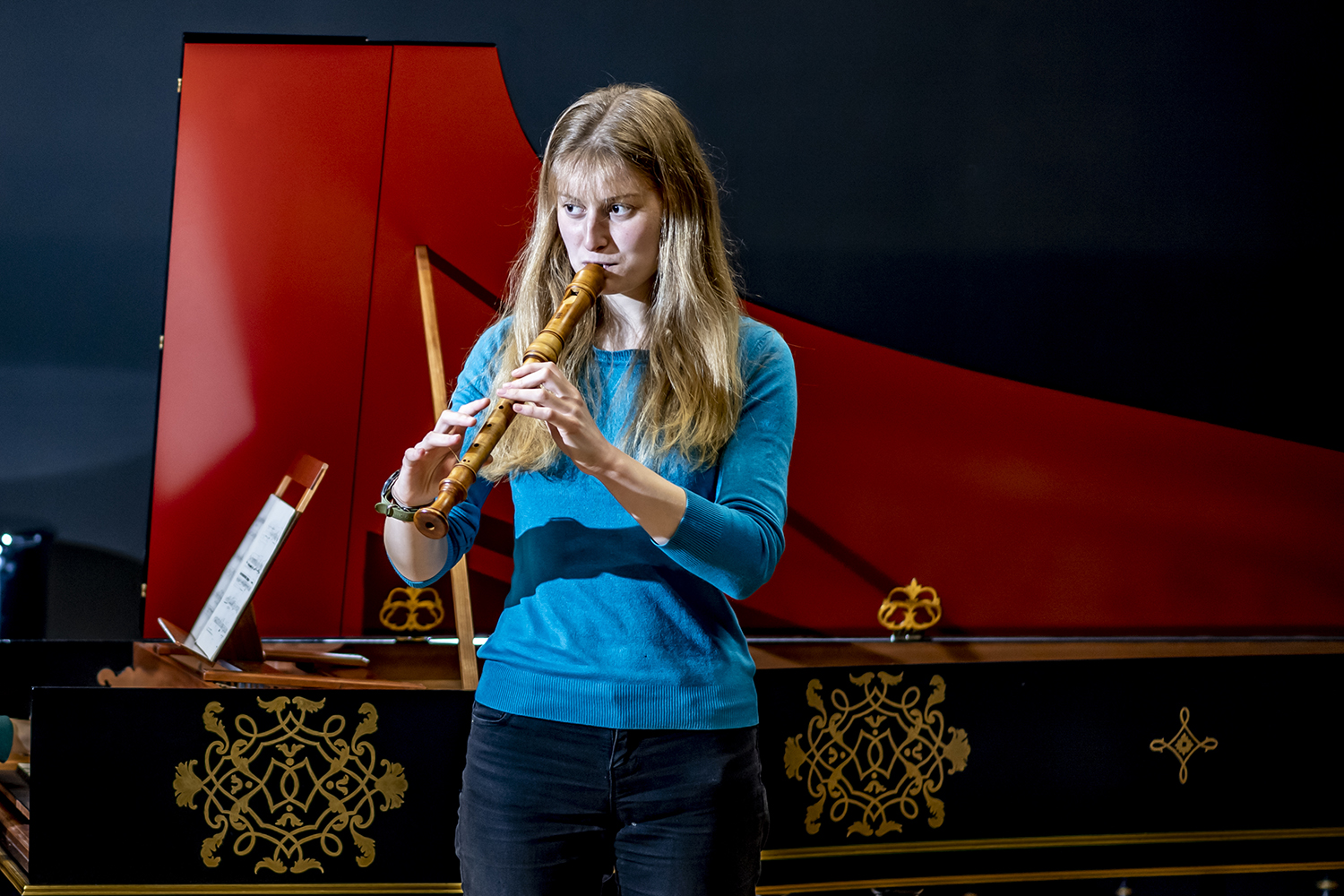 A woman with blonde hair and a blue jumper performing on a historical recorder in front of a harpsichord