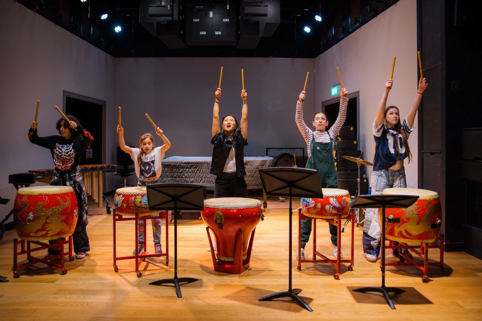 A group of people of various ages stand in front of drums raising mallets in the air