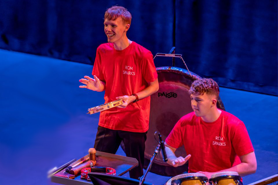 Two young men wearing red tshirts play percussion instruments on stage, smiling