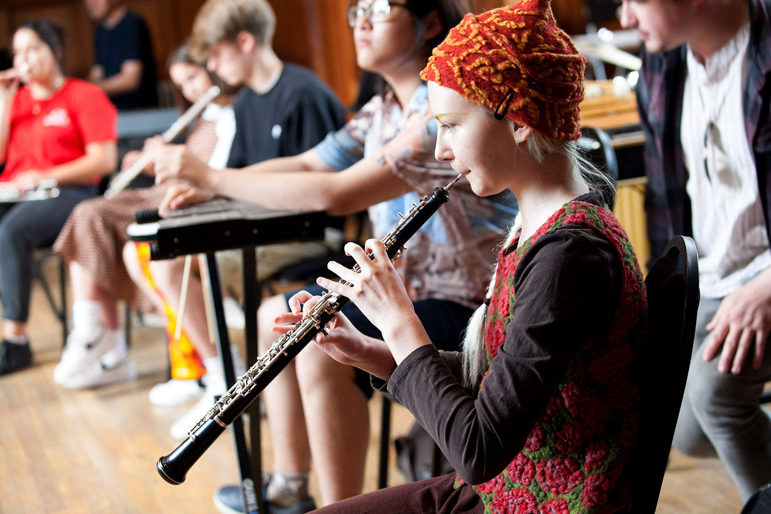 A young musician wearing a hat plays an oboe with other young musicians playing different instruments in the background