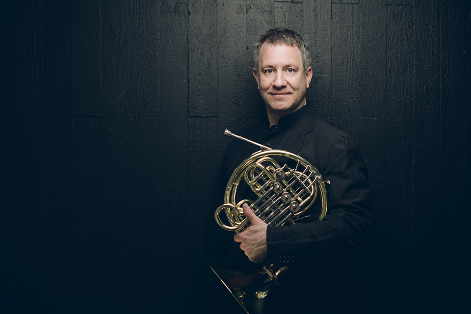 Stefan Dohr, wearing black, holds his horn under his arm, standing against a black background