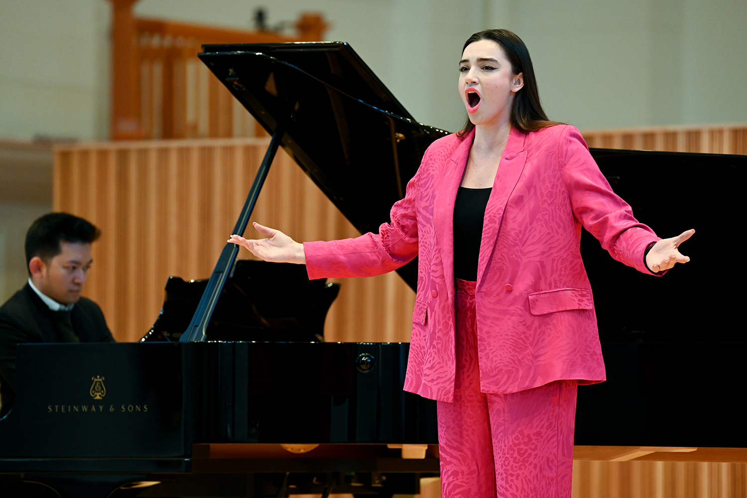 A woman wearing a pink suit sings on stage with a pianist playing in the background