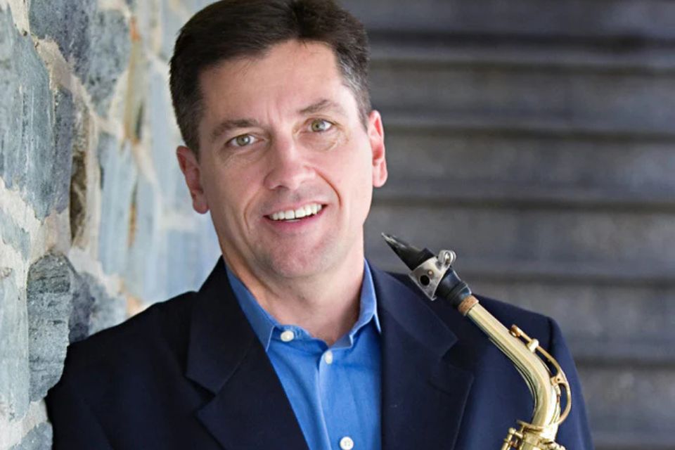 Timothy Roberts wears a blue shirt and black blazer and holds his saxophone, smiling