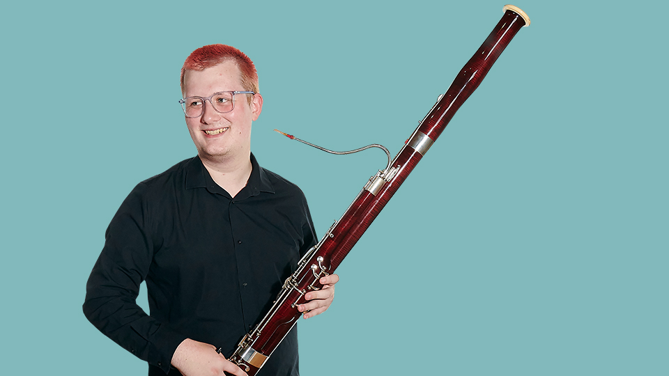 A male student wearing a black shirt, smiling, holding a bassoon.