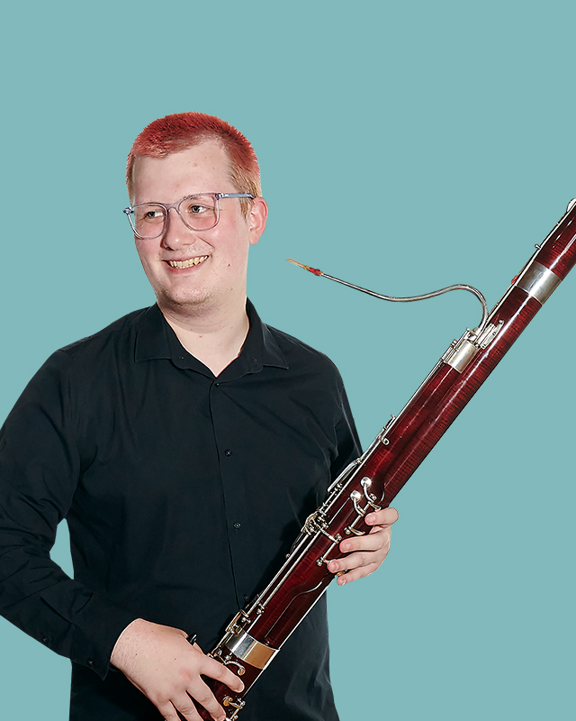 A male student wearing a black shirt, smiling, holding a bassoon.