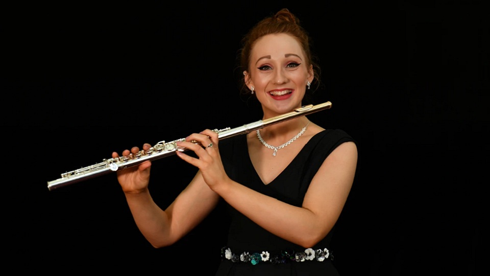 Photo of female alumna looking at the camera, wearing a black dress and holding a flute ready to play.