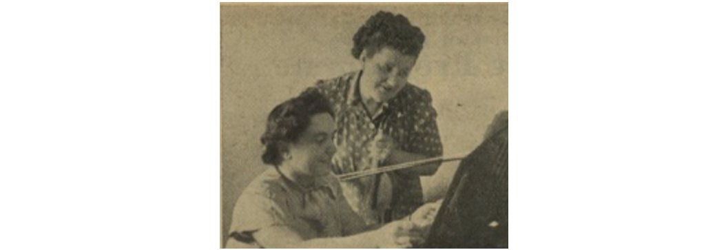 Two female musicians, with dark hair in braided hairstyles. One is sitting in front of a piano and the other is standing beside her; the one standing is holding a violin and both are looking at sheet music, wearing short sleeved dresses