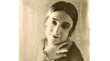 Slobadskaya’s press photo, a young woman is holding her left hand under her chin to pose with her head tilted to the side. She has dark hair which is tied back, she is wearing a lot of jewellery on her left wrist and hand.