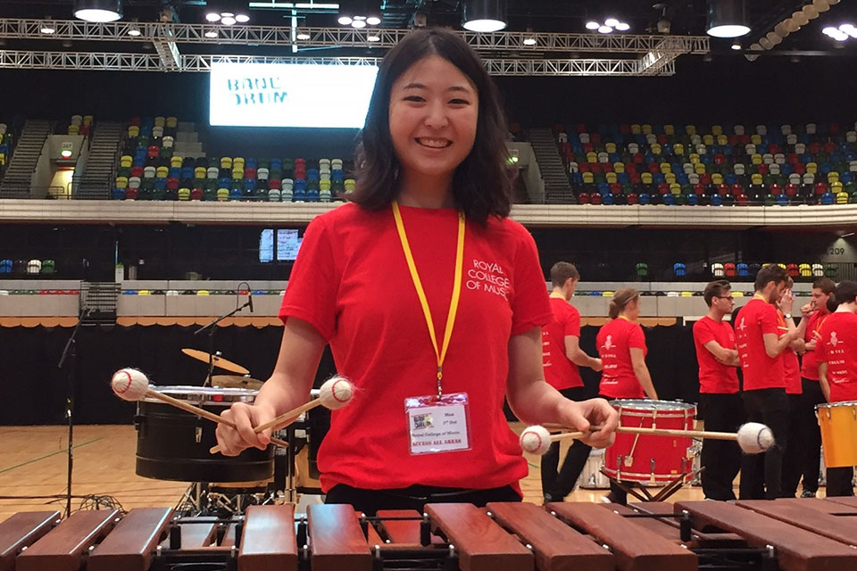 An RCM student smiling at the camera, playing the marimba, wearing a red RCM t-shirt