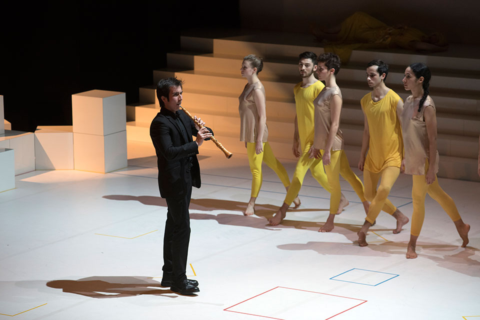 A male student performing a historical instrument on stage, with dancers in yellow clothing performing beside him.