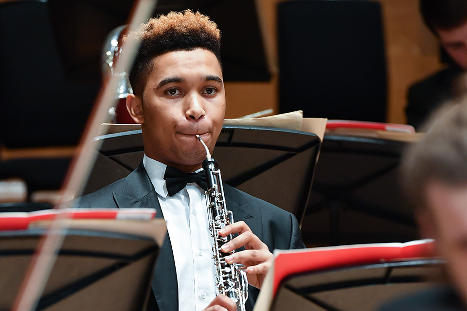 A black male student, dressed in smart clothes, performing on a clarinet, performing in an orchestra setting.