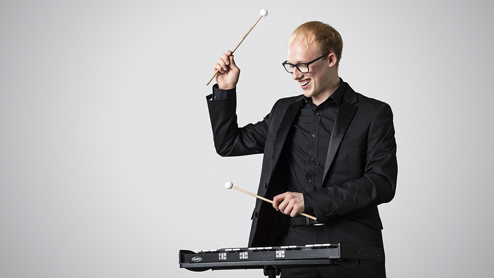 A student playing the xylophone smiling, against a grey background