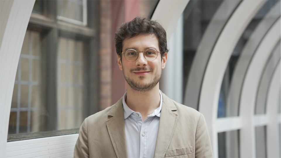 Headshot of a white man with curly brown hair and glasses, smiling at the camera in a well light hallway at the Royal College of Music.